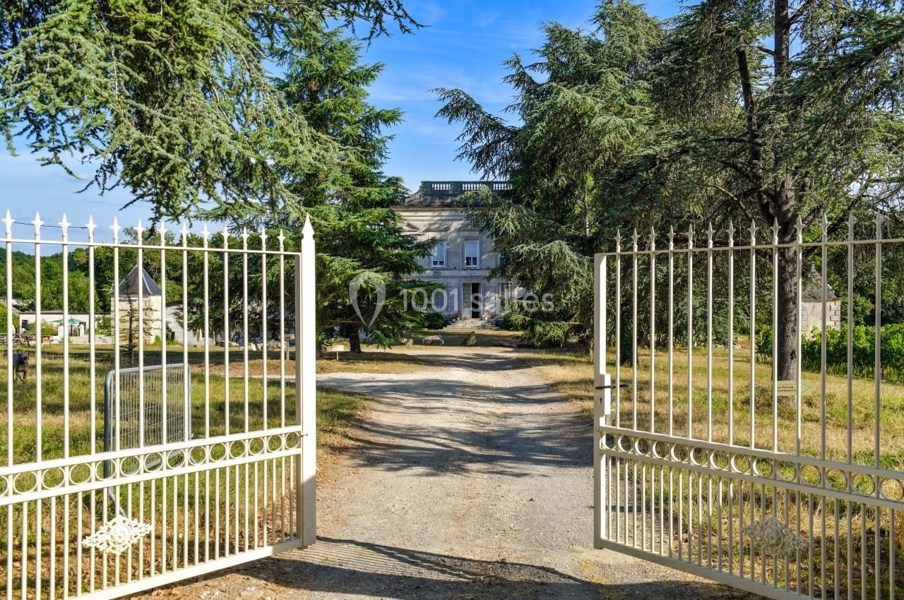 Portail en fer forgé ouvert menant à une allée bordée d'arbres et à un bâtiment en pierre sous un ciel dégagé.