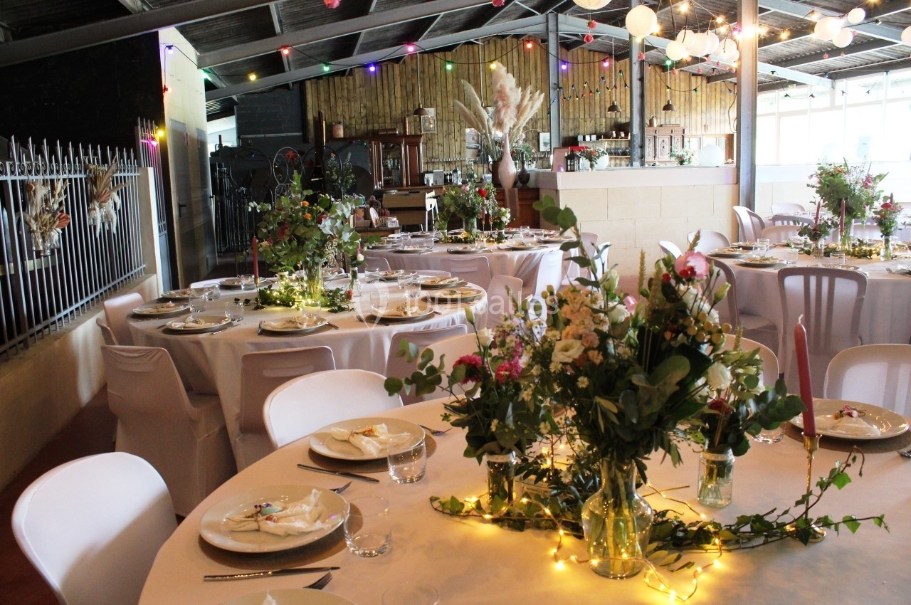 Salle décorée pour un événement avec tables rondes dressées, guirlandes lumineuses et arrangements floraux.