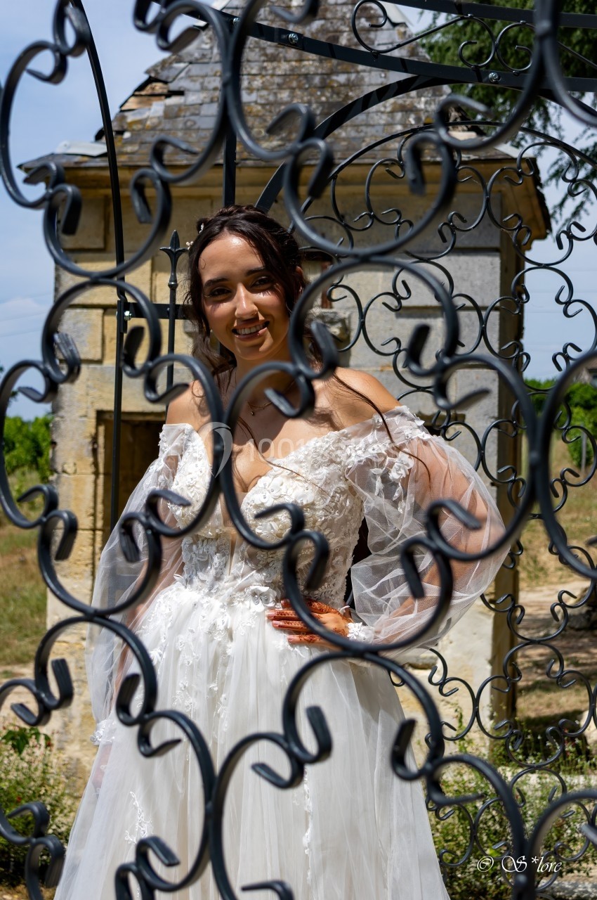 Femme en robe de mariée blanche posant derrière une grille en fer forgé, avec un bâtiment en pierre en arrière-plan.