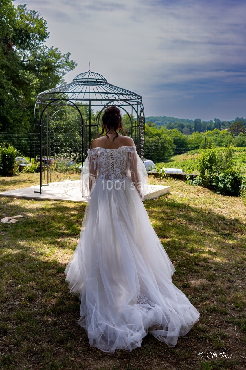 Une femme en robe de mariée blanche marche vers une tonnelle métallique dans un jardin verdoyant.