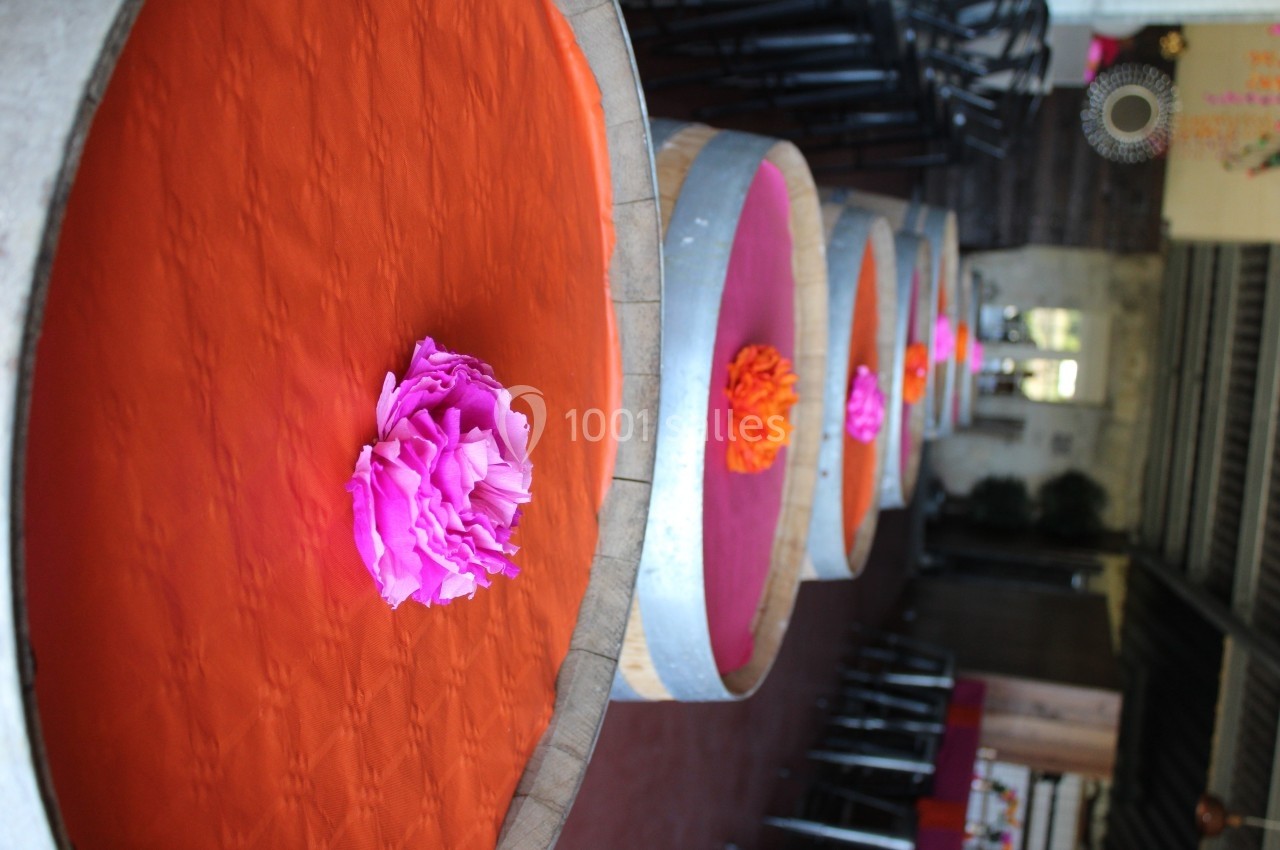 Tables rondes recouvertes de tissus orange avec des fleurs en papier rose, alignées dans une salle intérieure.