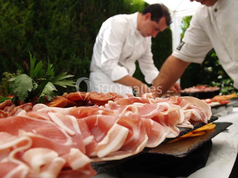 Plateau de charcuterie avec des tranches de jambon et saucisson, préparé par deux chefs en arrière-plan.