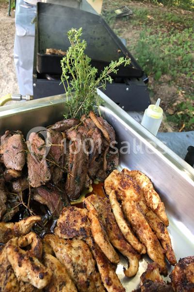 Viandes grillées variées dans un plat en métal, avec des herbes fraîches, devant un barbecue en plein air.