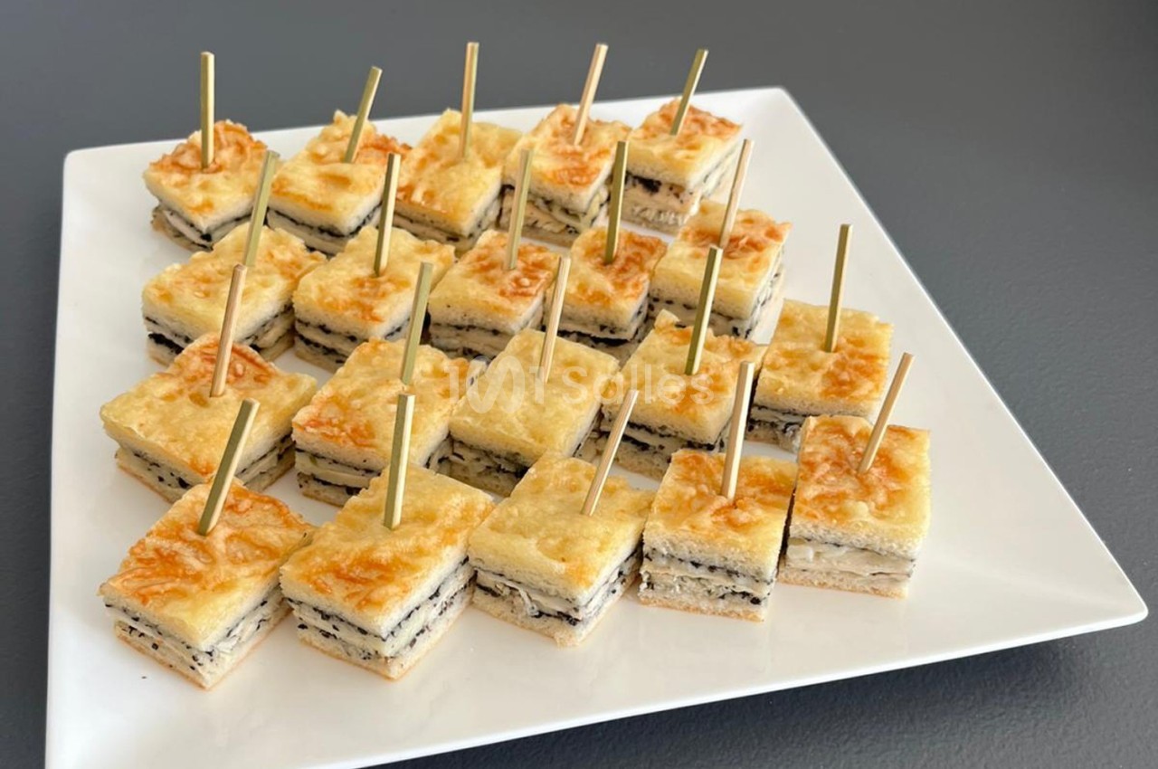 Carrés de croque-monsieur garnis de fromage et truffe, présentés sur un plateau blanc avec des piques en bois.