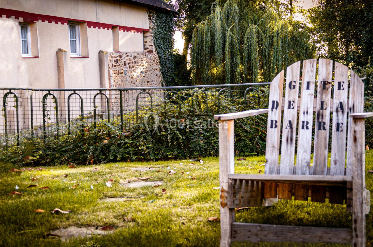 Chaise en bois marquée ’Gîte de la Barre’ dans un jardin verdoyant près d'une maison en pierre et crépi.