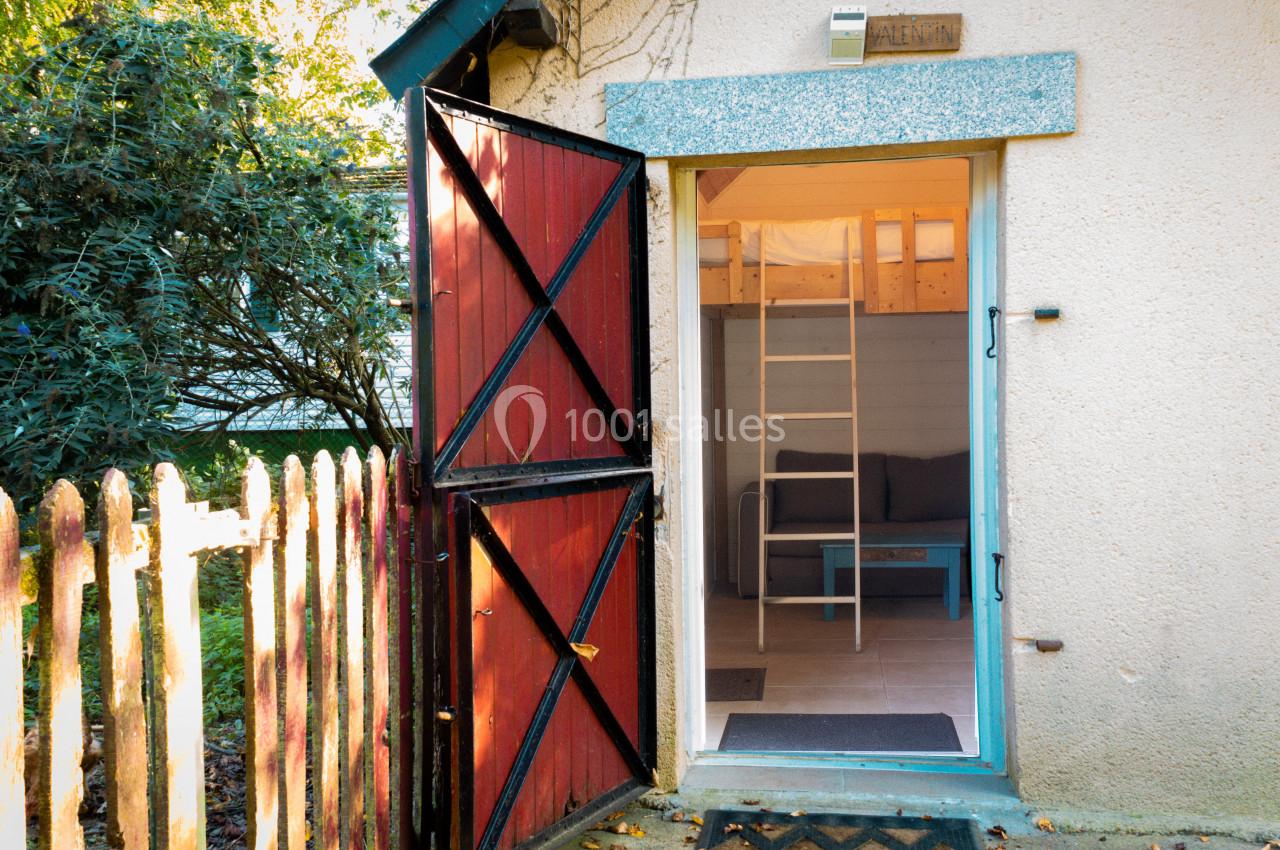 Porte rouge ouverte donnant sur l'entrée d'une petite maison avec un intérieur lumineux et un canapé visible.