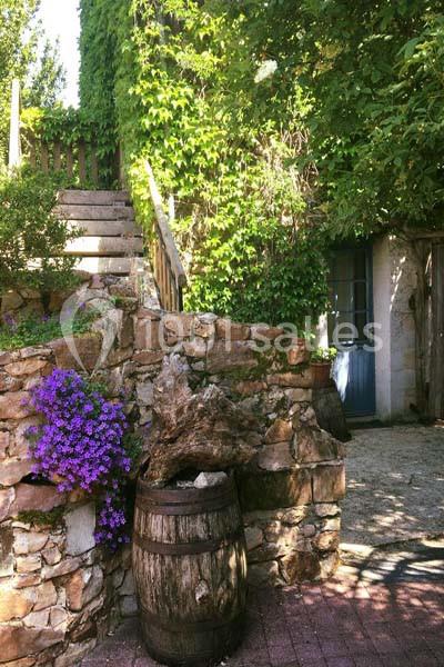 Escalier en pierre bordé de végétation, tonneau en bois et fleurs violettes devant une maison avec porte bleue.
