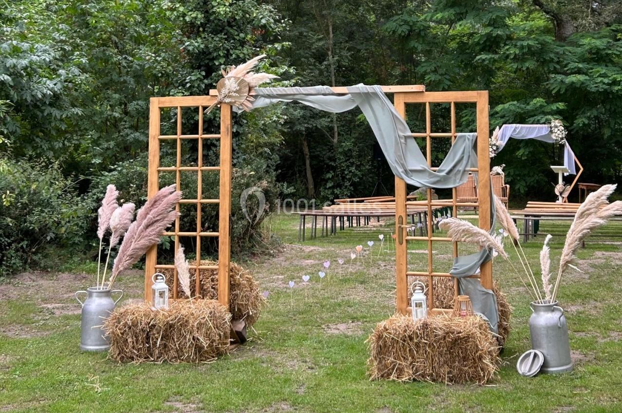 Arche en bois décorée de voilages, pampas et lanternes, installée dans un jardin pour une cérémonie en plein air.