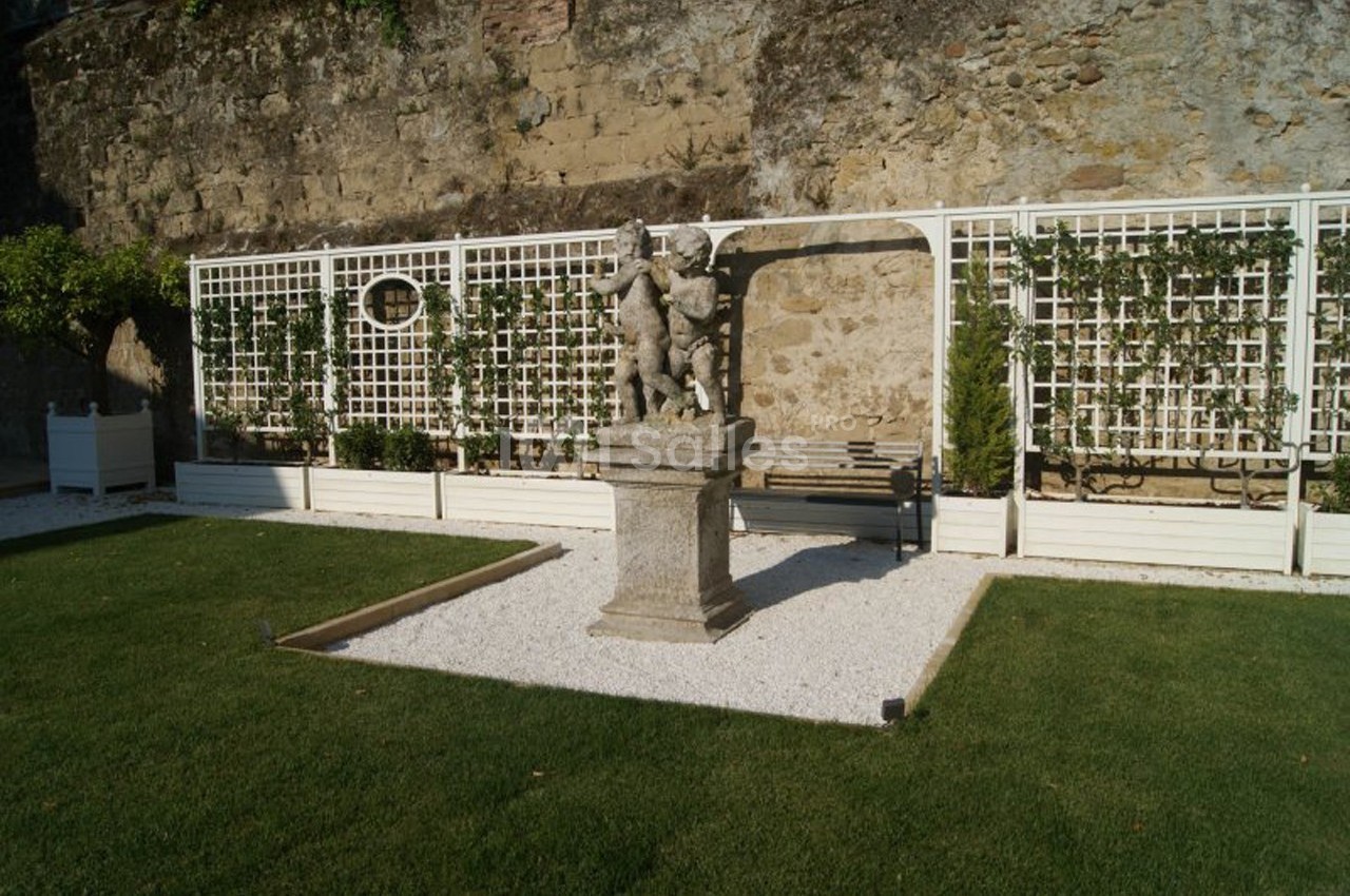 Statue en pierre sur un socle, entourée de gravier, devant une treille et un mur en pierre dans un jardin aménagé.
