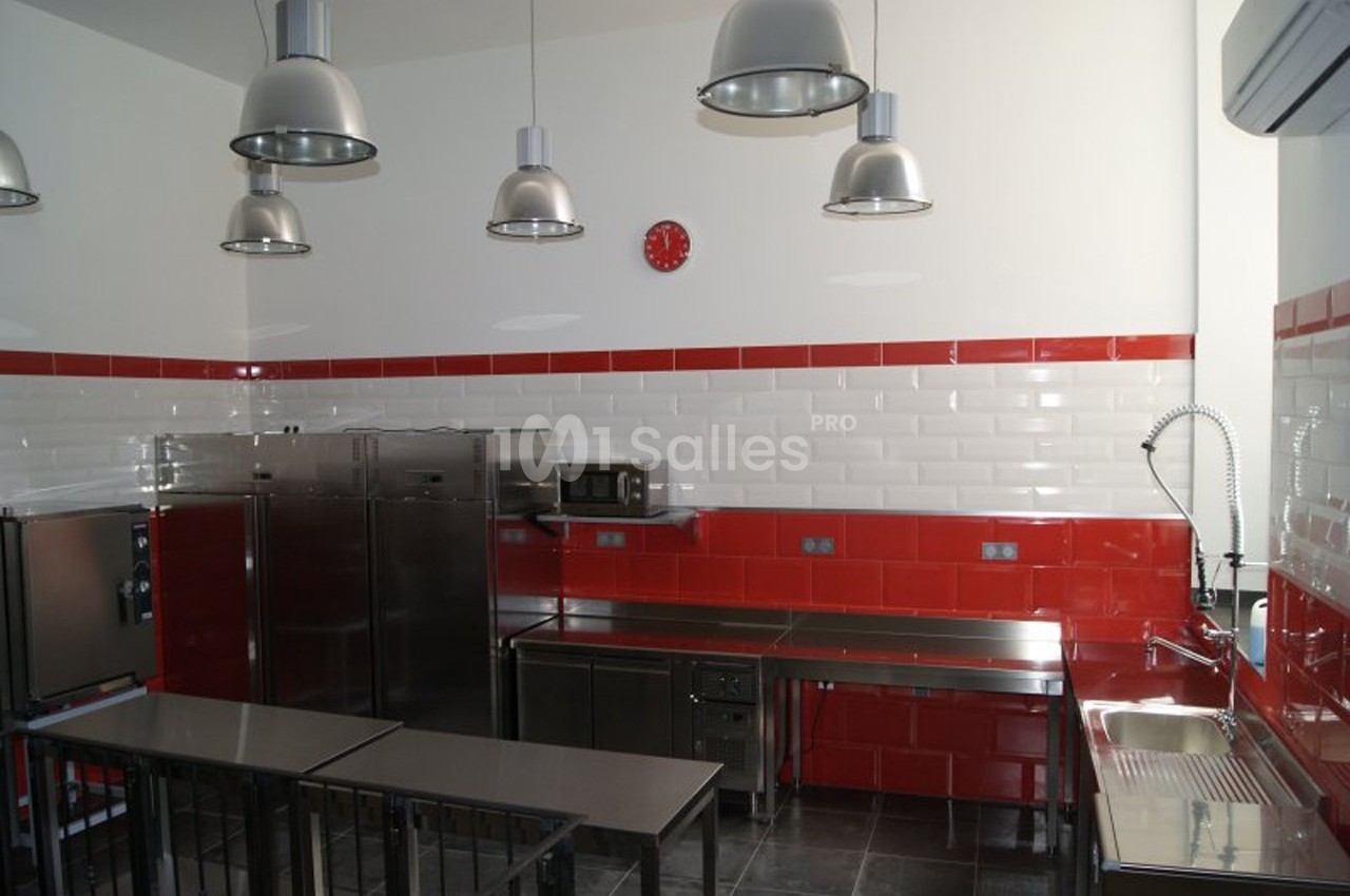 Cuisine professionnelle avec mobilier en inox, murs carrelés rouges et blancs, éclairage suspendu et horloge murale rouge.