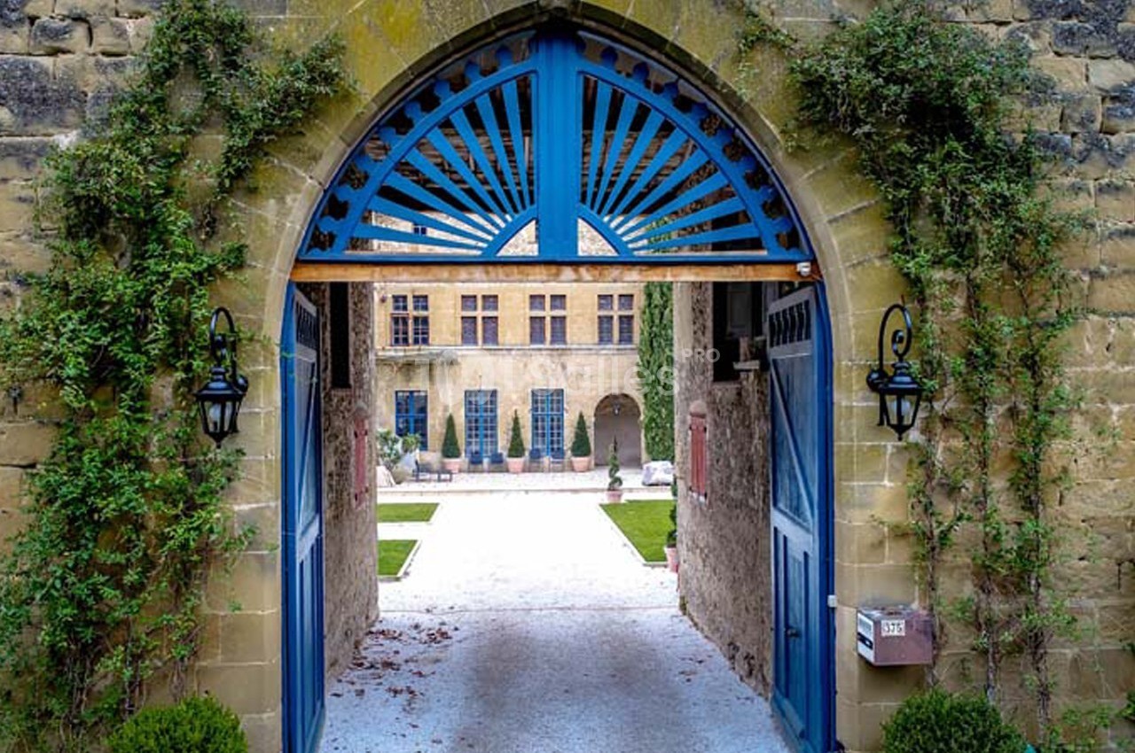 Vue d'une cour intérieure à travers une arche en pierre ornée de portes bleues et entourée de végétation.