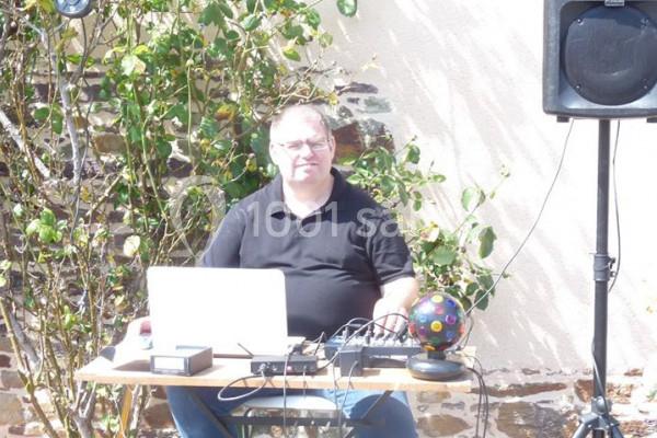 Homme assis devant un ordinateur portable et du matériel audio, installé à l'extérieur près d'un mur clair et de végétation.
