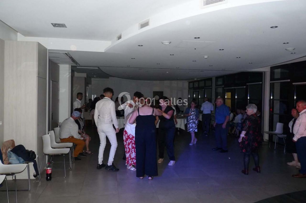 Des personnes dansent et discutent dans une salle éclairée, avec des chaises alignées contre les murs.