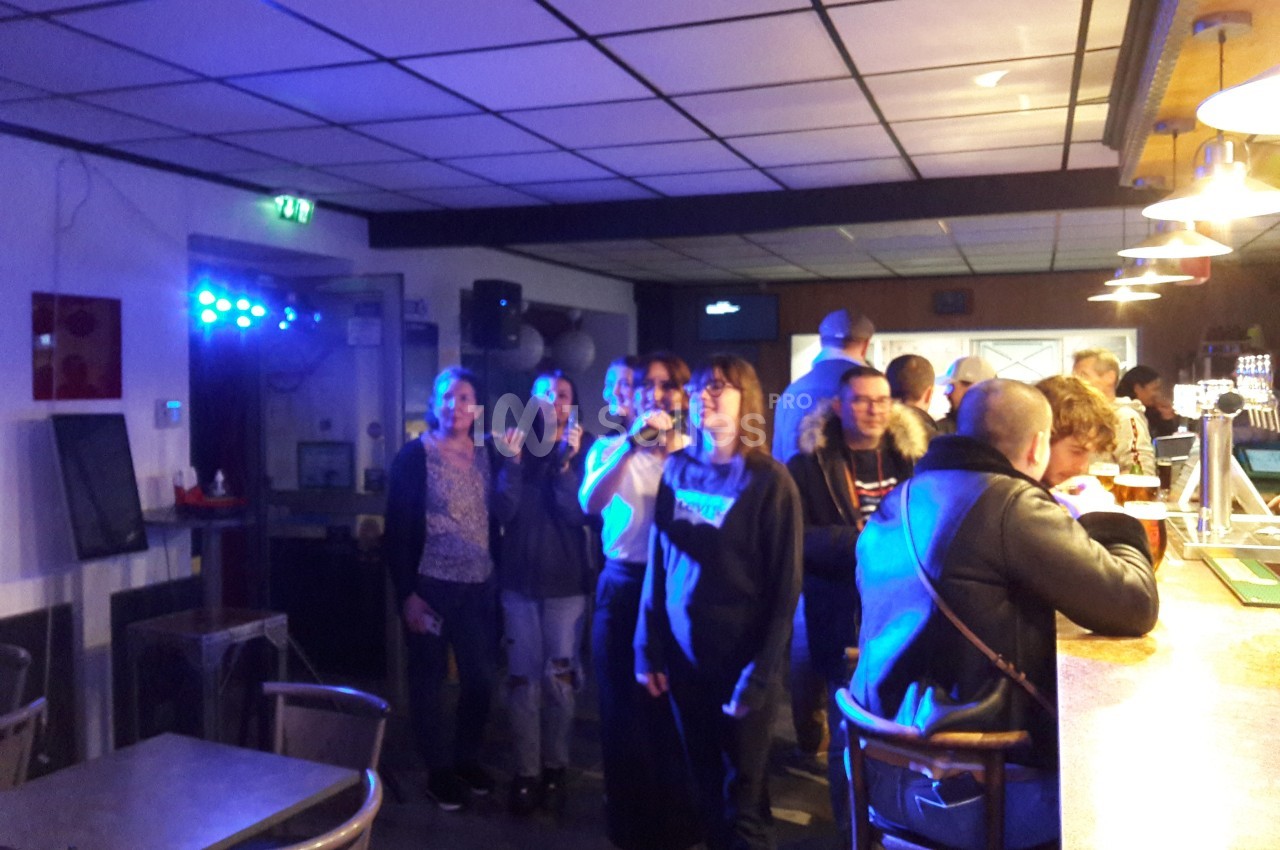 Des personnes debout dans un bar, certaines sourient face à l'objectif, éclairées par une lumière bleutée.