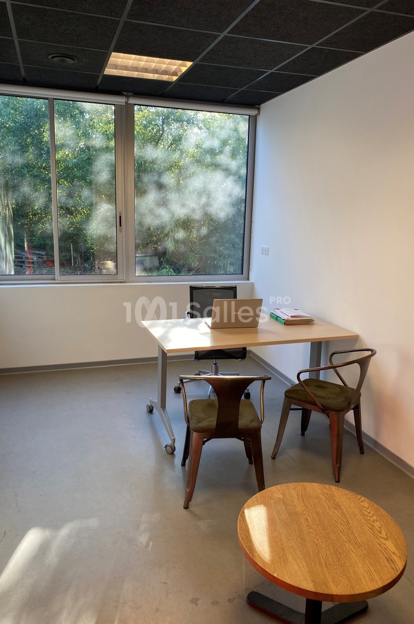 Bureau lumineux avec une table, deux chaises, un ordinateur portable et une fenêtre donnant sur un espace arboré.