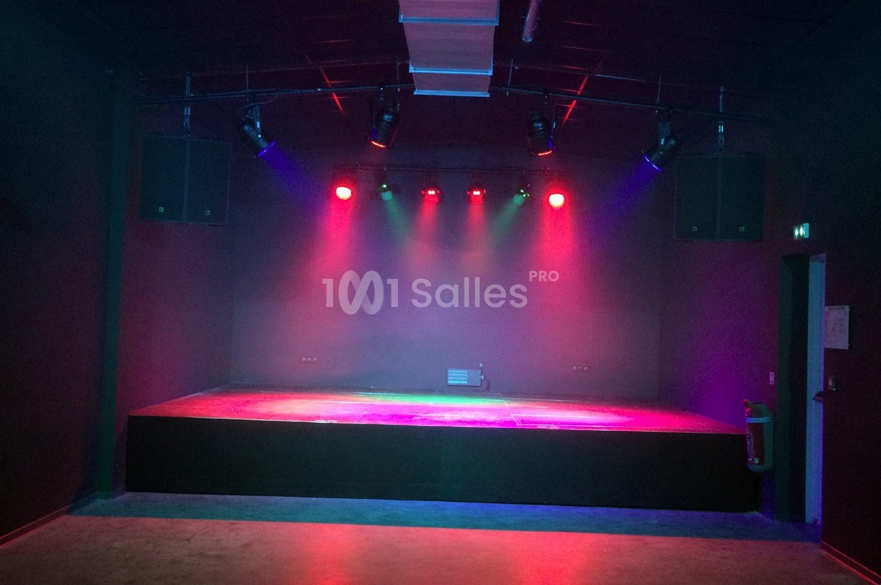 Scène vide éclairée par des projecteurs colorés dans une salle sombre avec un sol en béton.