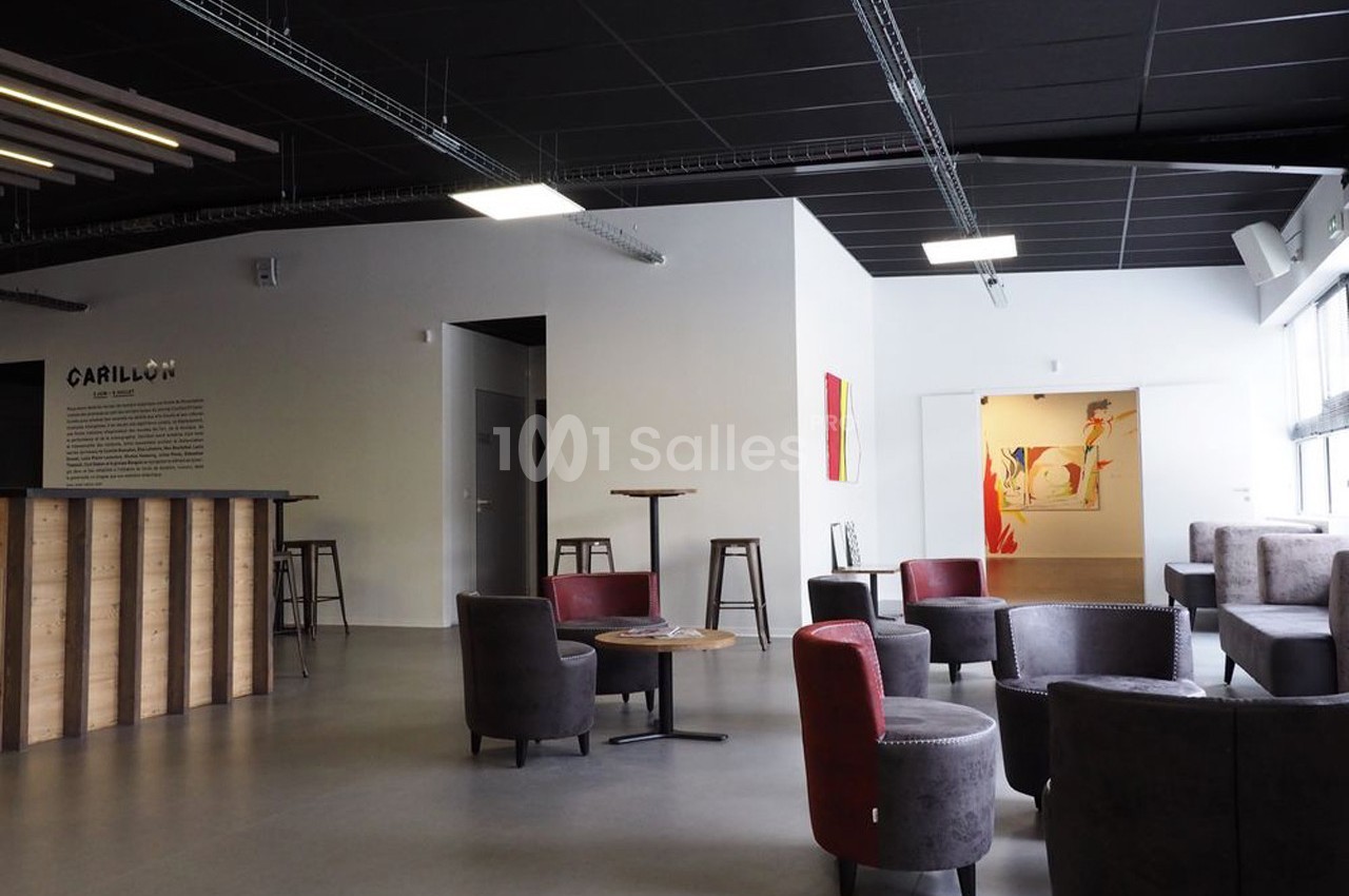 Espace lounge moderne avec fauteuils, tables basses, comptoir en bois et murs décorés, éclairé par des fenêtres.