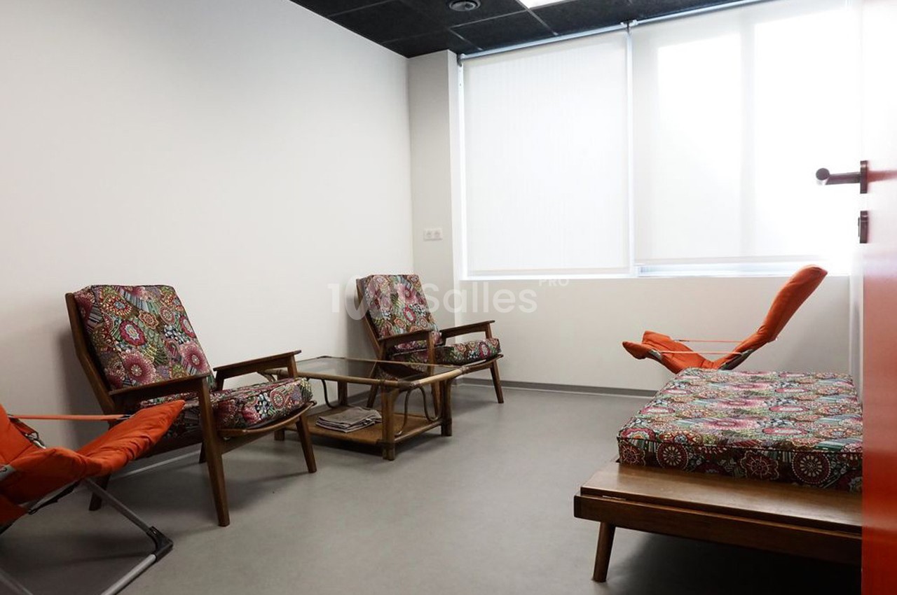 Salle lumineuse avec fauteuils et banquette en bois, coussins colorés, table basse en verre et fenêtre avec store blanc.