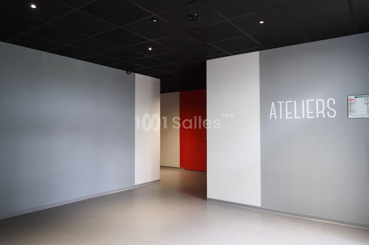Mur gris avec l'inscription ’ATELIERS’, sol clair, plafond noir et porte rouge au fond d'un couloir.