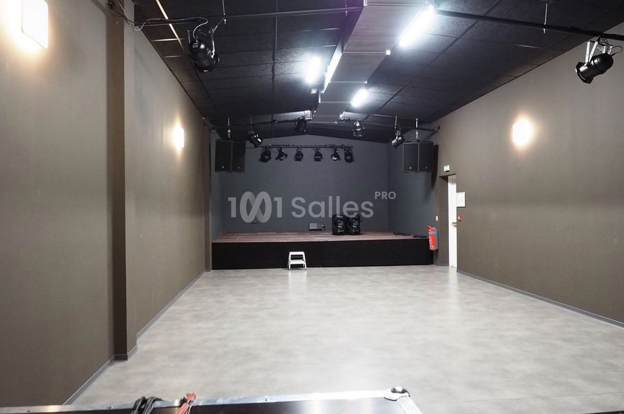 Salle vide avec sol en béton, murs sombres, éclairage au plafond et petite scène surélevée au fond.