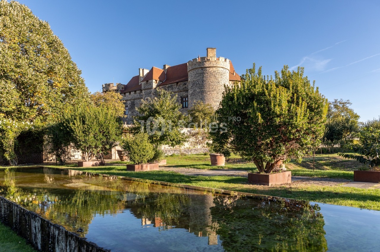 Vue d'un château en pierre entouré d'arbres, avec un bassin réfléchissant le bâtiment et le ciel bleu.