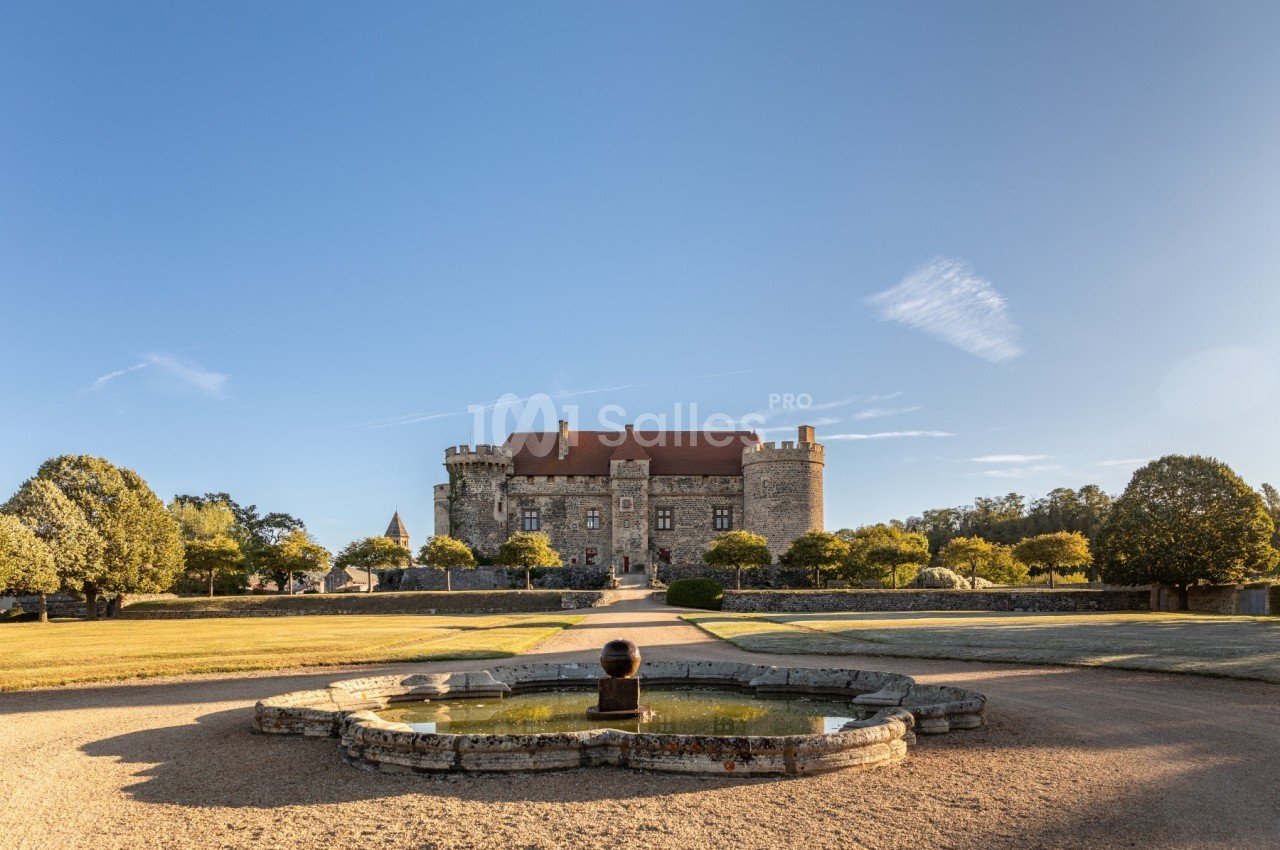 Vue d'un château en pierre avec un toit rouge, entouré de jardins et d'une fontaine au premier plan.