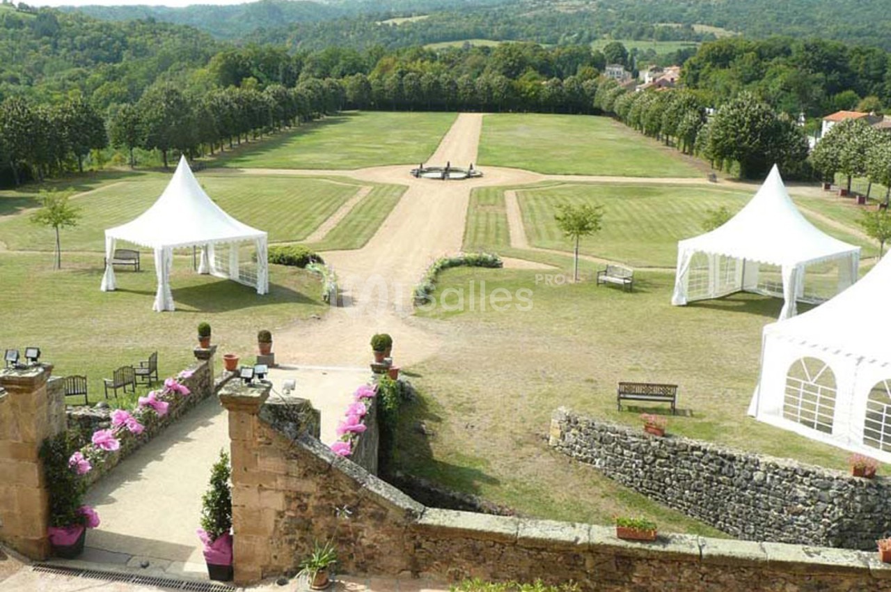 Vue d'un grand jardin avec allées bordées d'arbres, pelouses, tentes blanches et collines en arrière-plan.