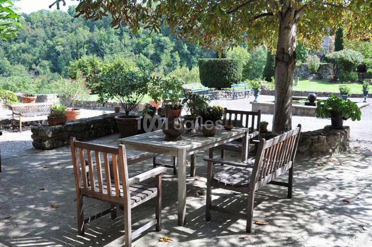 Table et chaises en bois disposées sous un arbre dans un jardin verdoyant avec des pots de fleurs et une vue sur la nature.