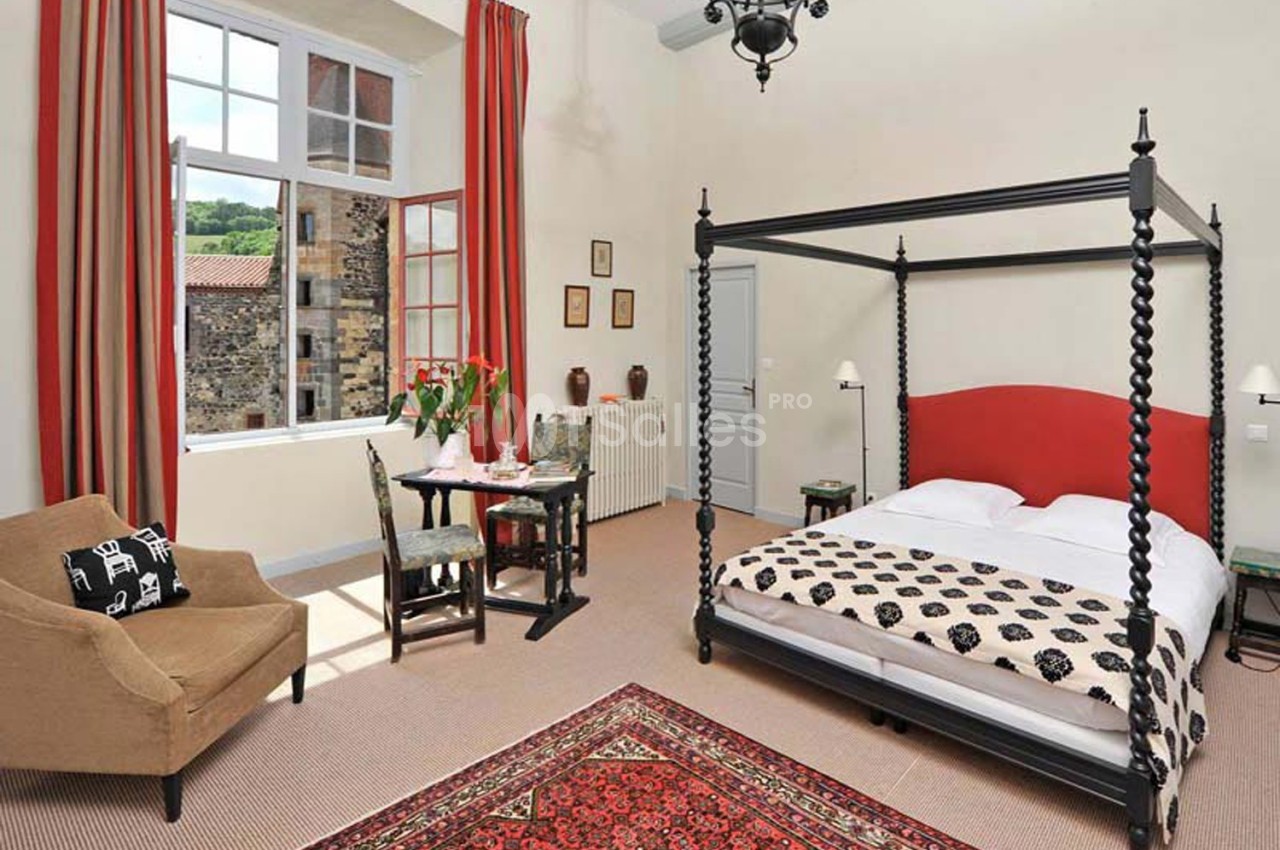 Chambre lumineuse avec lit à baldaquin, fauteuil, table dressée, tapis rouge et grande fenêtre donnant sur l'extérieur.