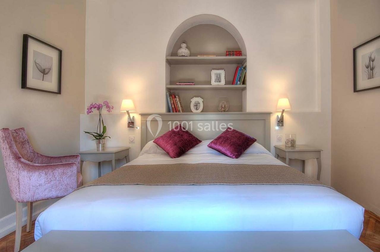 Lit double avec coussins violets, tables de chevet, étagère murale décorée et fauteuil rose dans une chambre lumineuse.
