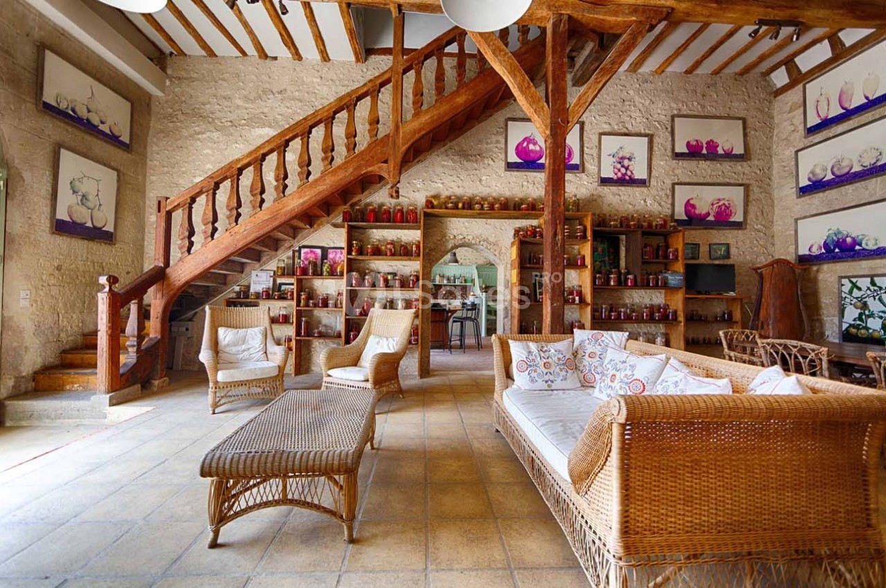 Salon rustique avec mobilier en rotin, escalier en bois, murs en pierre et étagères décorées de pots et cadres colorés.