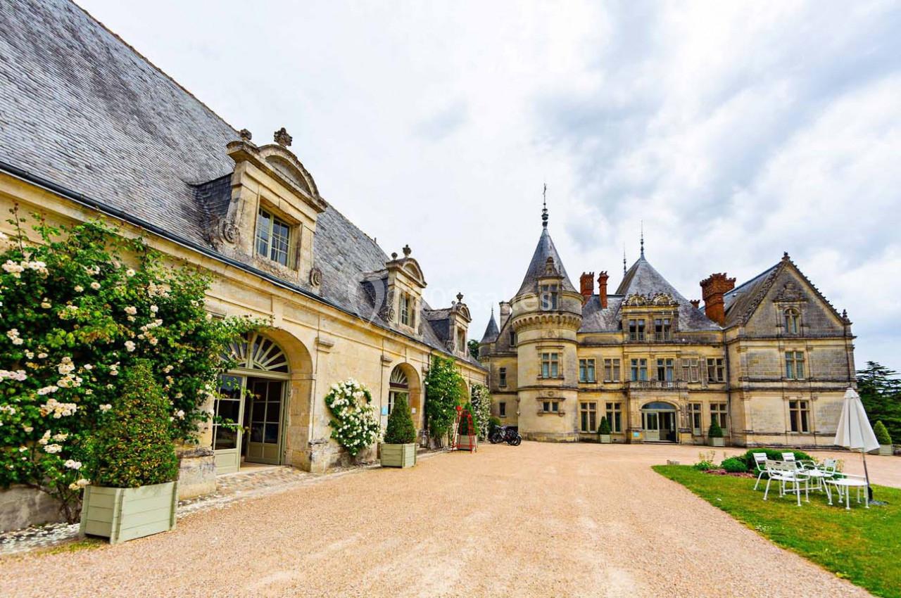 Façade d'un château historique avec tours, jardin fleuri et mobilier d'extérieur sur une allée gravillonnée.