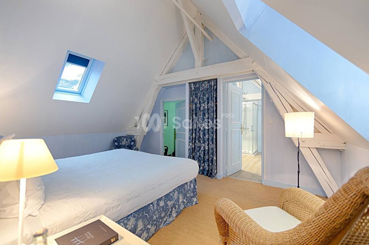 Chambre mansardée lumineuse avec lit double, fauteuil en osier, lampes et accès à une salle de bain adjacente.