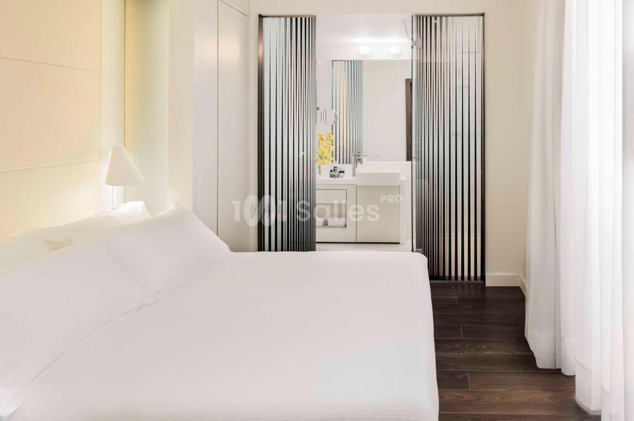 Chambre lumineuse avec lit double, parquet foncé et salle de bain attenante séparée par des panneaux coulissants.