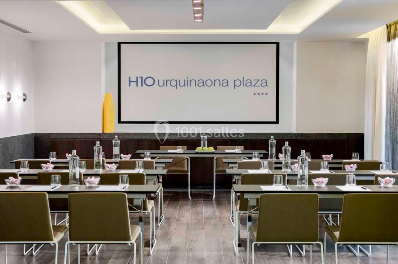 Salle de réunion lumineuse avec tables alignées, chaises, écran de projection et bouteilles d'eau disposées.