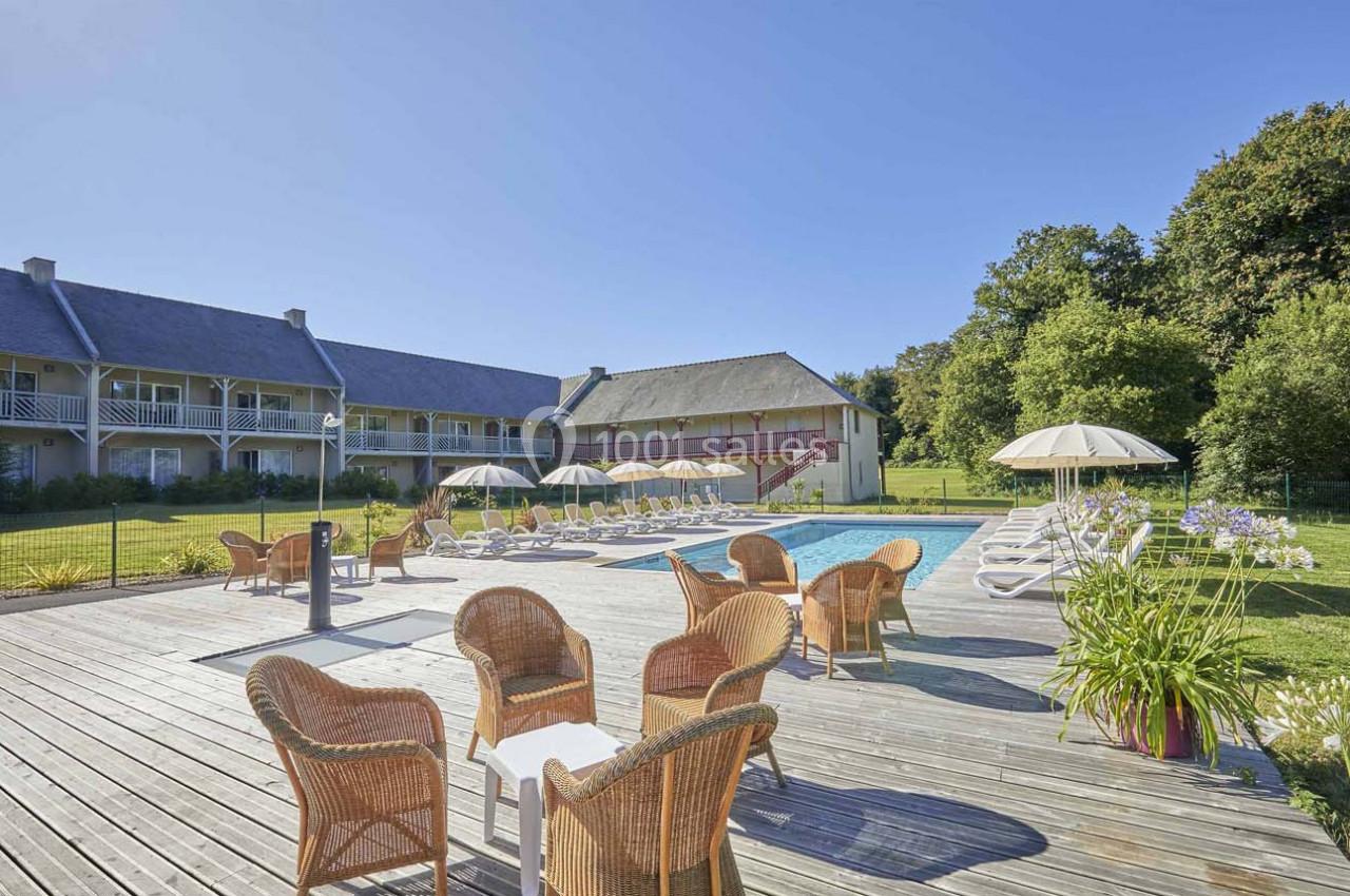 Terrasse en bois avec fauteuils en rotin, piscine extérieure entourée de transats et parasols, bâtiments en arrière-plan.