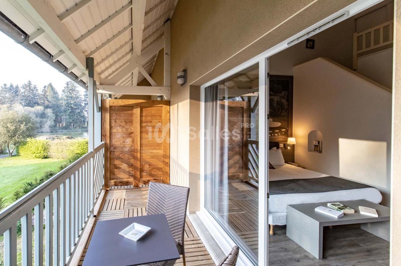 Vue d'une chambre d'hôtel avec balcon, table et chaises, donnant sur un paysage verdoyant et un plan d'eau.