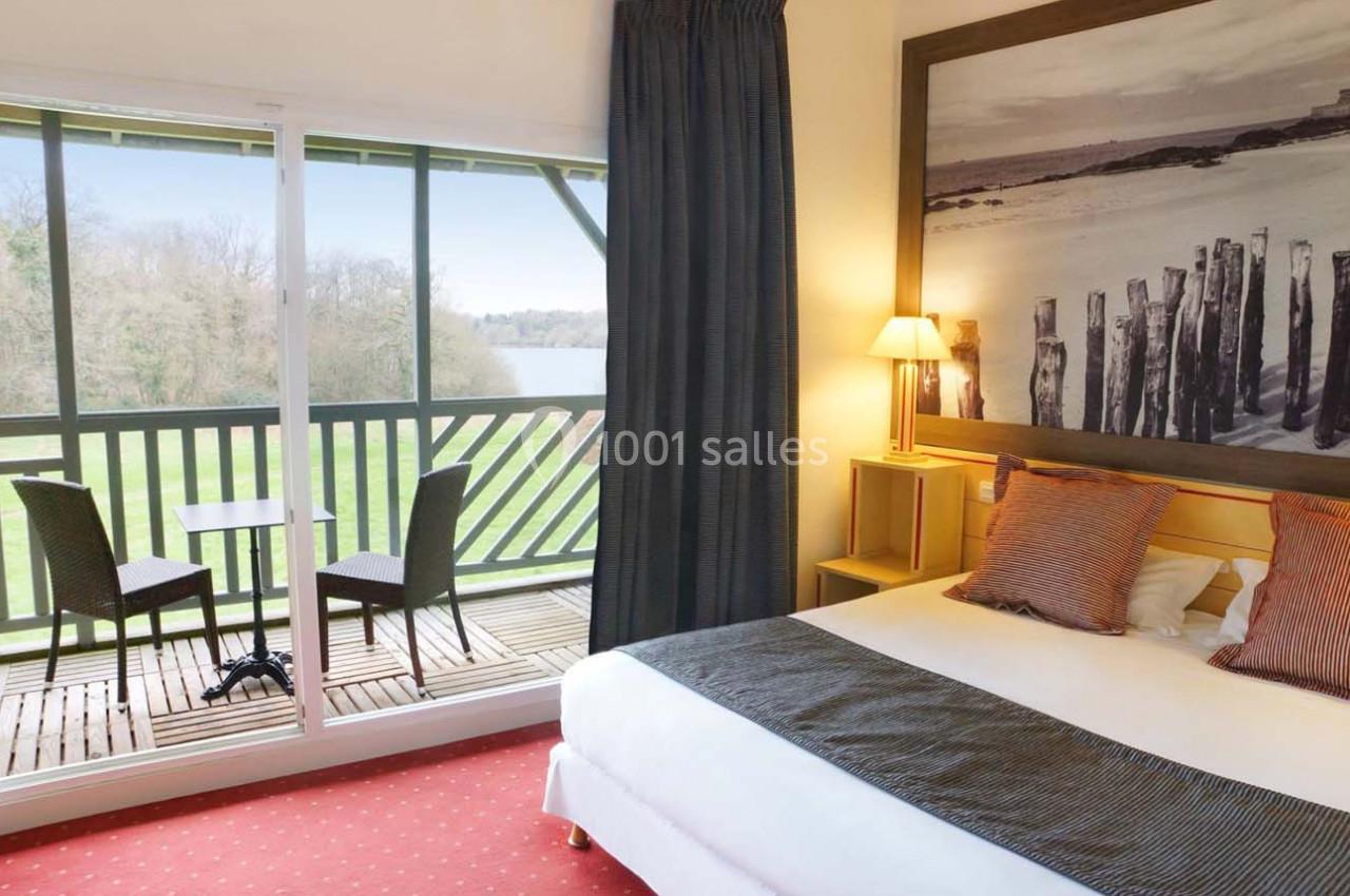 Chambre lumineuse avec lit double, balcon aménagé, vue sur un paysage verdoyant et une étendue d'eau.