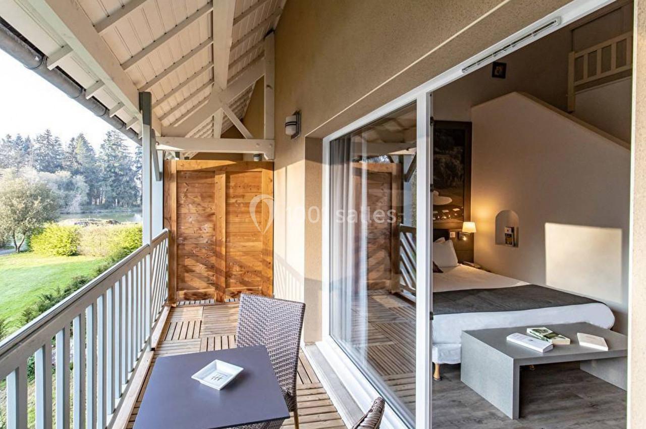 Balcon en bois avec table et chaises, vue sur un espace vert, donnant sur une chambre lumineuse avec un lit.