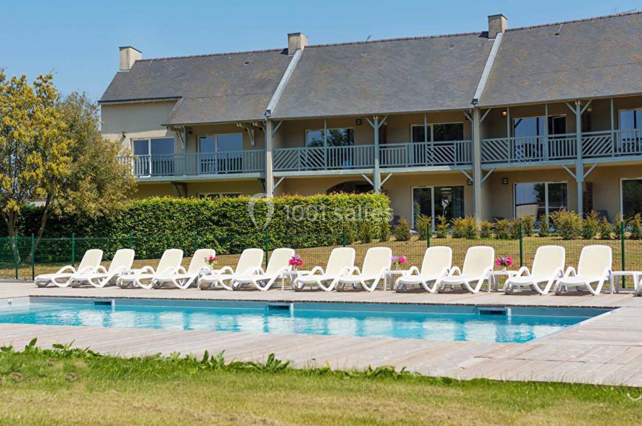 Piscine extérieure entourée de transats blancs devant un bâtiment à deux étages avec balcons en bois.