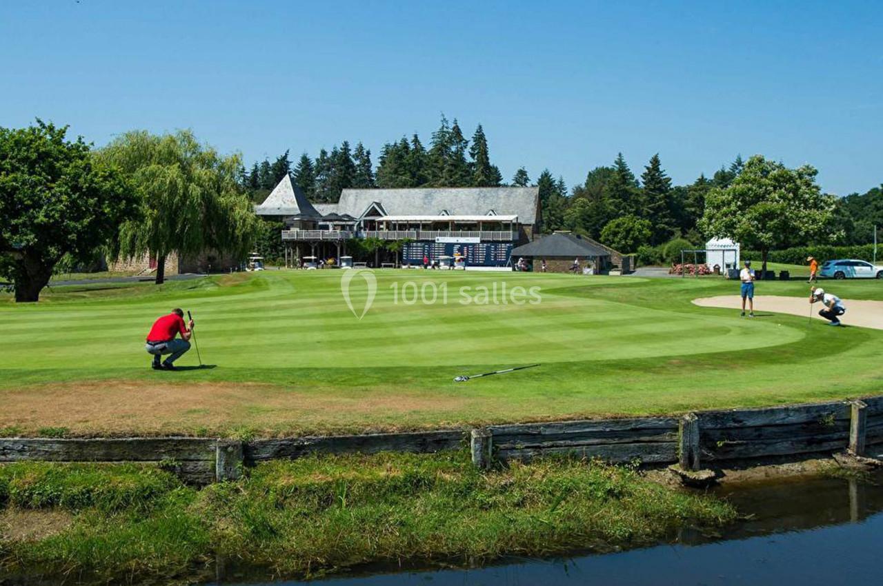 Un golfeur s'apprête à putter sur un green entouré de végétation, avec un club-house en arrière-plan.
