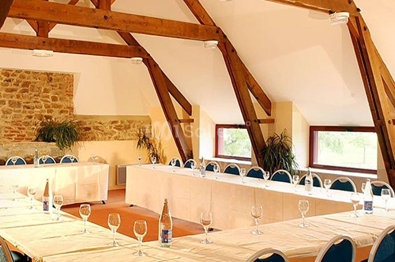 Salle de réunion lumineuse avec poutres apparentes, tables en U dressées avec verres et chaises bleues.