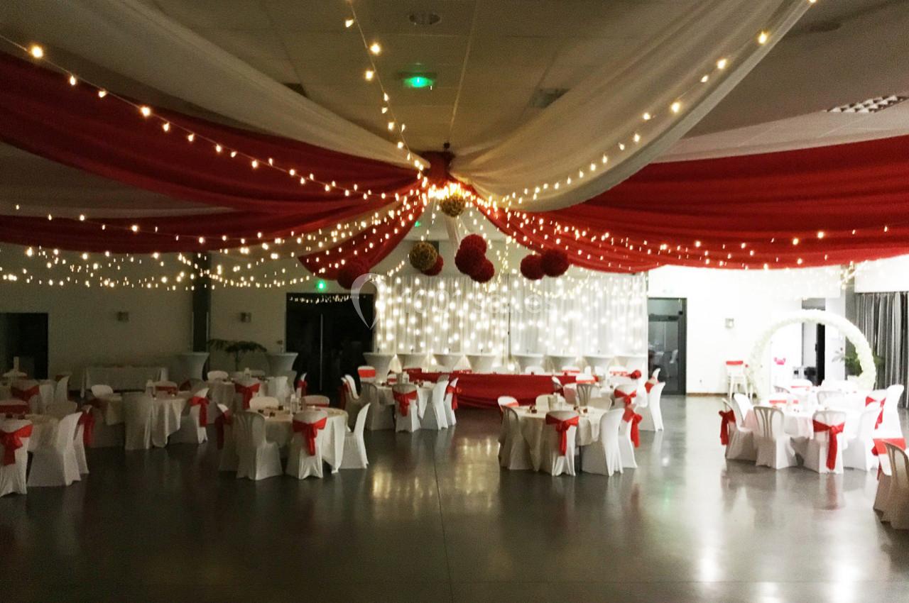 Salle de réception décorée de drapés rouges et blancs, guirlandes lumineuses et tables dressées avec chaises ornées de nœuds…