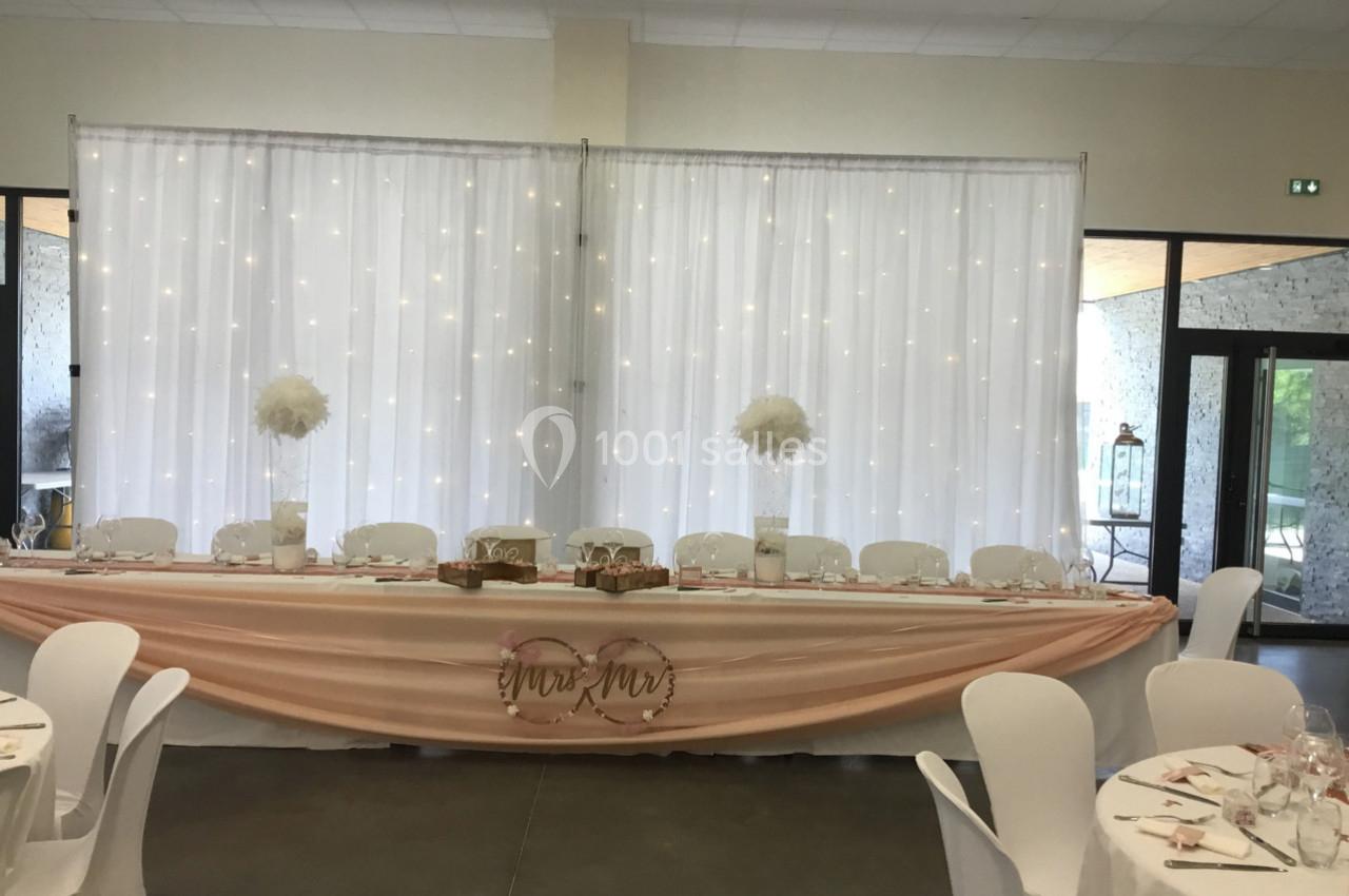 Table d'honneur décorée avec nappes roses, fleurs blanches et rideau lumineux en arrière-plan dans une salle de réception.