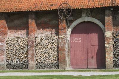 Miniature Location salle Clairmarais (Pas-de-Calais) - La Ferme de l'Abbaye #18 Façade d'une grange en briques avec porte en bois rouge, toit en tuiles et piles de bois empilées sur les côtés.