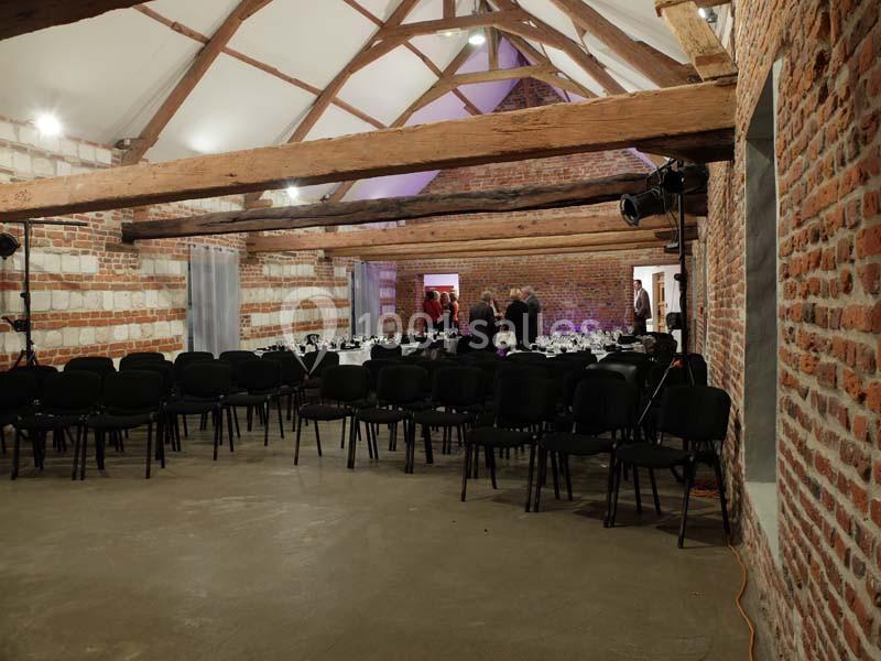 Salle en briques avec poutres apparentes, rangées de chaises noires et tables dressées pour un événement.