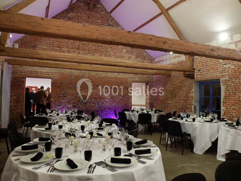 Salle en briques apparentes avec des tables rondes dressées pour un événement, éclairage tamisé et poutres en bois visibles.