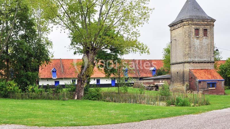 Vue d'une ferme traditionnelle avec bâtiments en briques, toit rouge, tour en pierre et jardin verdoyant.