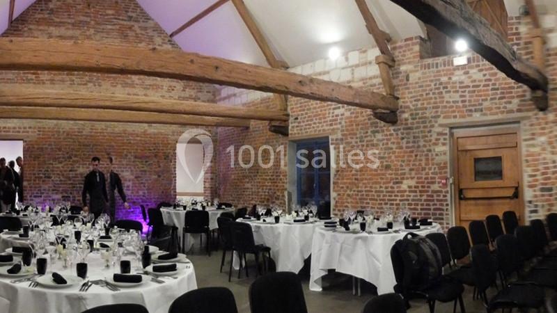 Salle en briques avec poutres apparentes, tables rondes dressées pour un événement, chaises noires et lumière tamisée.