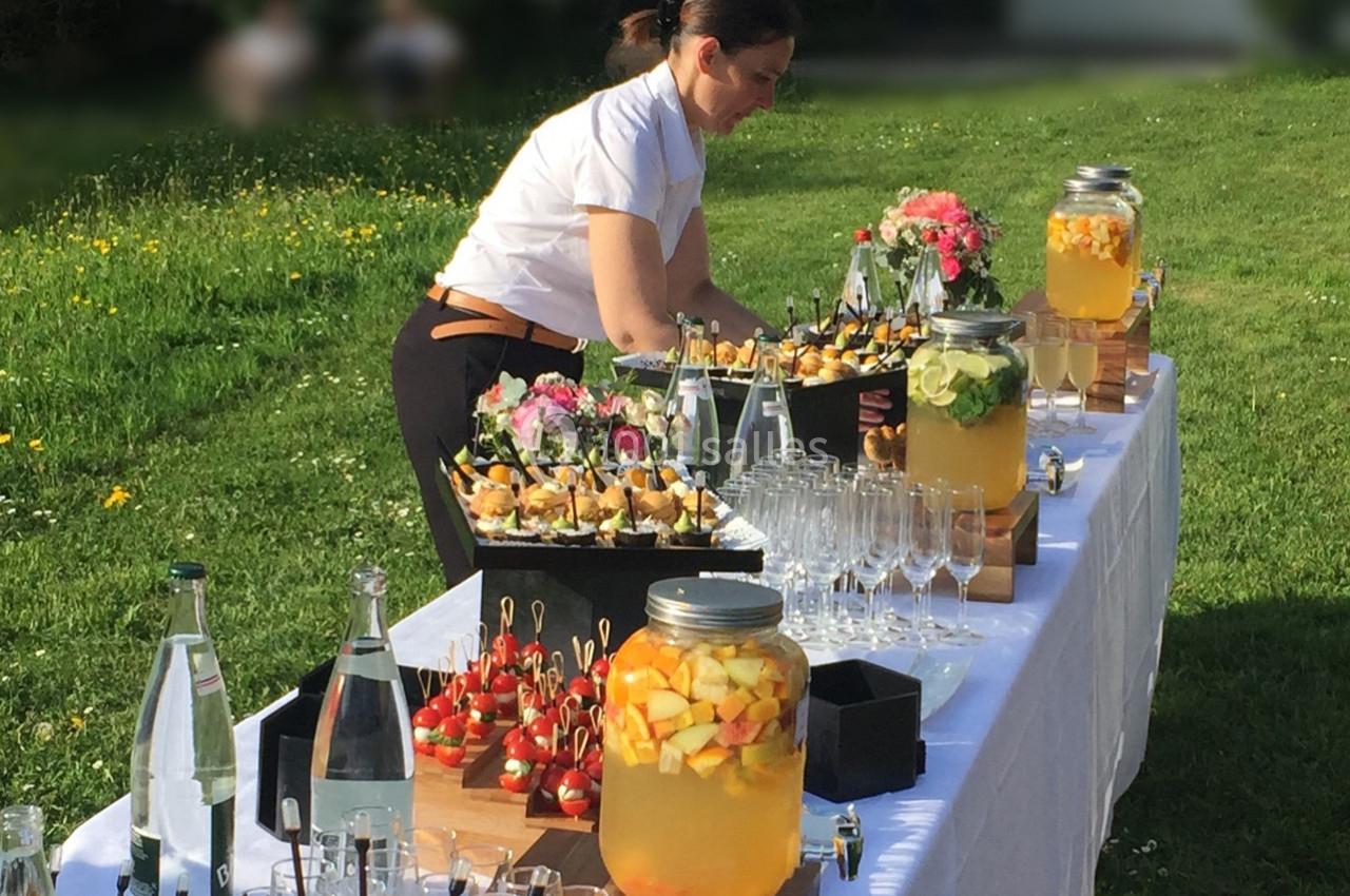 Une femme prépare un buffet en plein air avec boissons, verrines et décorations florales sur une table blanche.